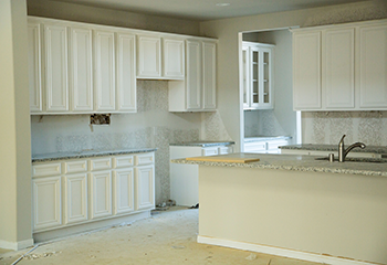 The image shows a newly remodeled kitchen with freshly installed white cabinets, a large kitchen island with a sink, and new countertops. The space is bright and modern, showcasing the quality of the remodeling work.